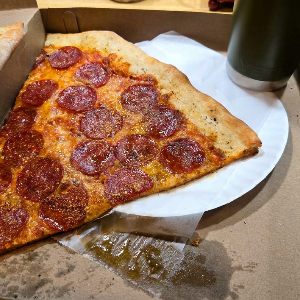 Large pepperoni pizza slice in a cardboard box beside another slice and a dark drink on a wooden table.